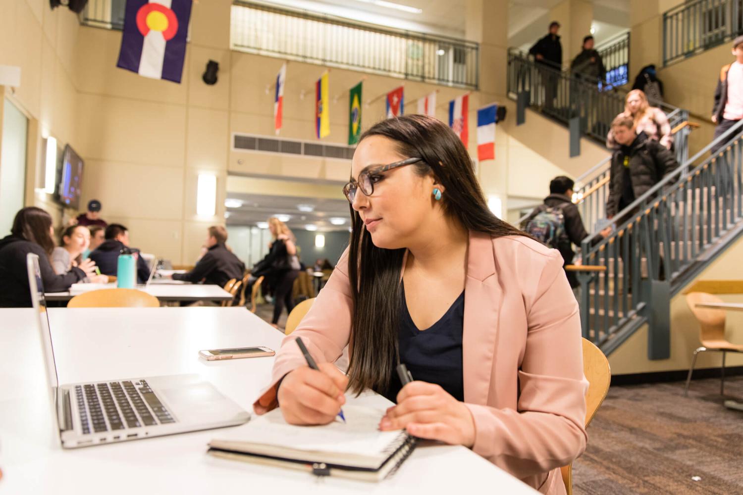 A student studies on her laptop in the common area of the international building on campus with students moving in the background