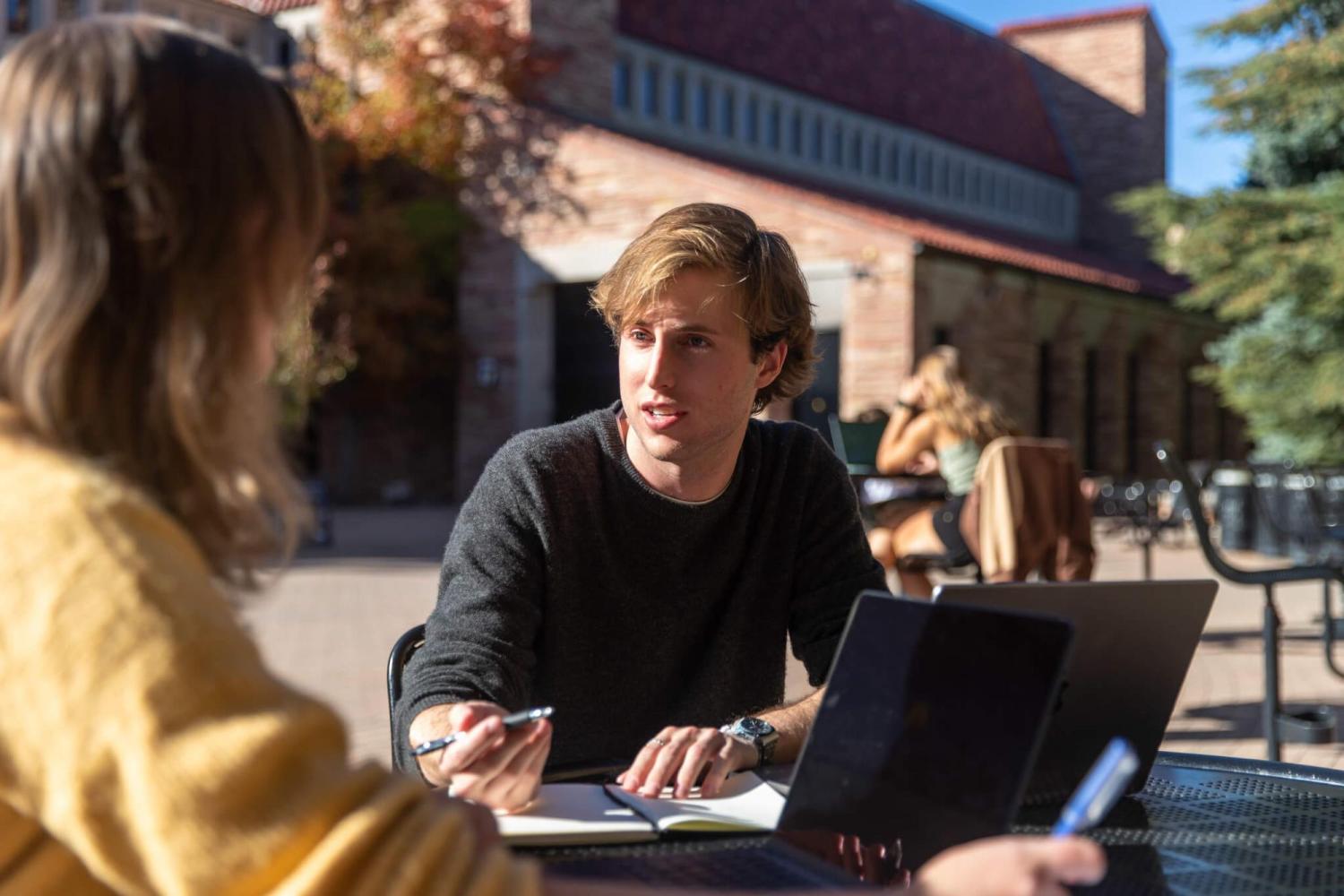 Two students talk together at an outdoor table on campus while using their laptops and writing on their notepads