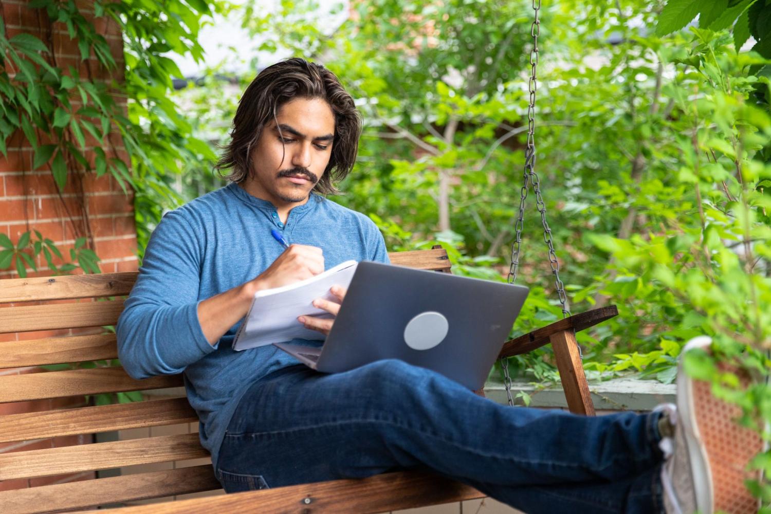 A student takes notes on a note pad while working on his laptop on a porch swing