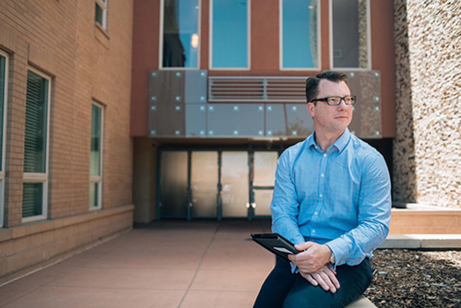 Man in blue button-down shirt sitting on concrete planter box in front of building